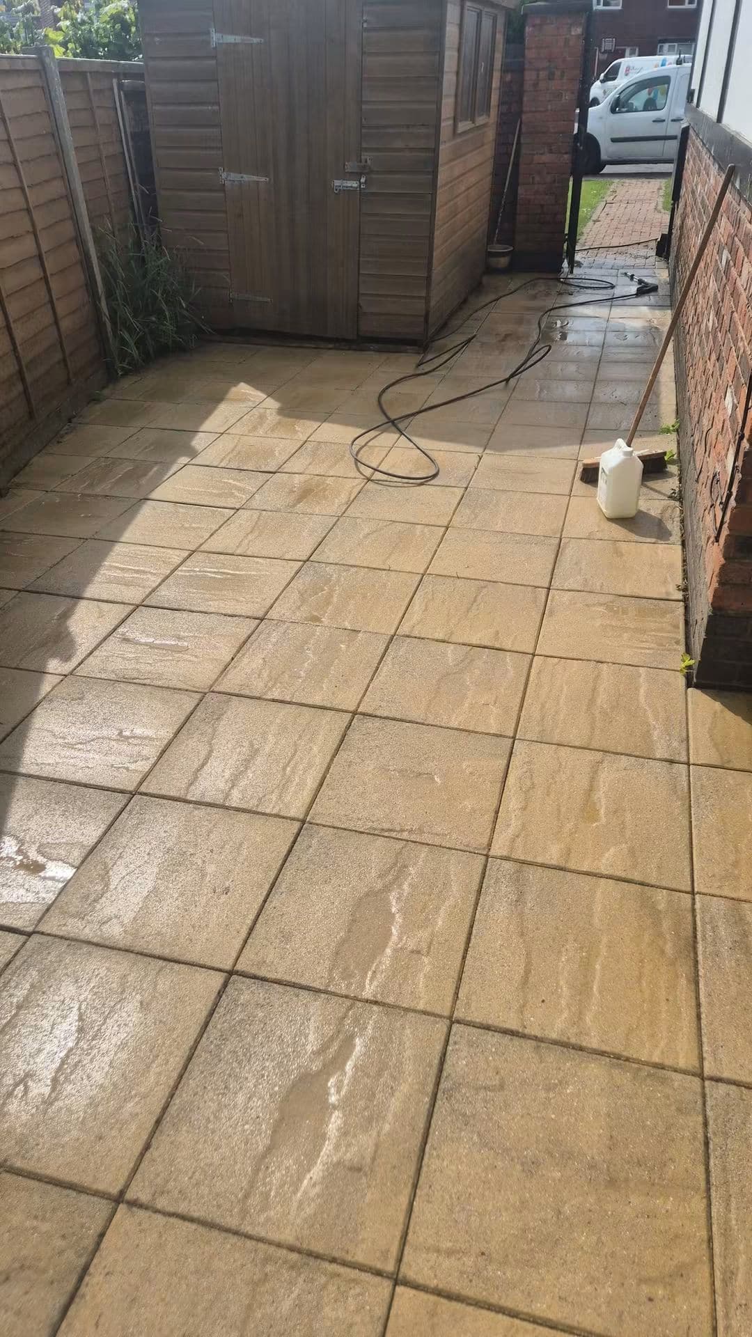 Golden paving slabs in a side garden being pressure washed, with hose, lance and brush on the patio